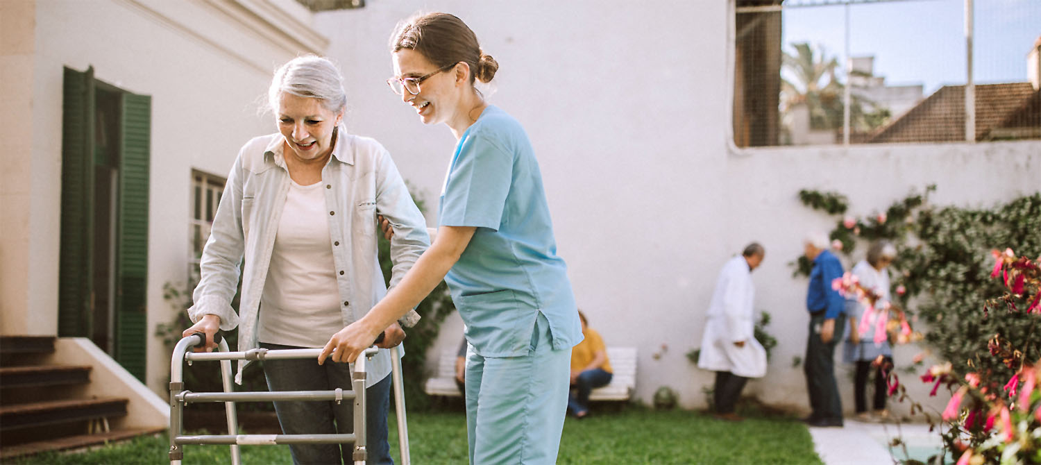 Photo of nursing assistant helping a woman with a walker outside in a courtyard.